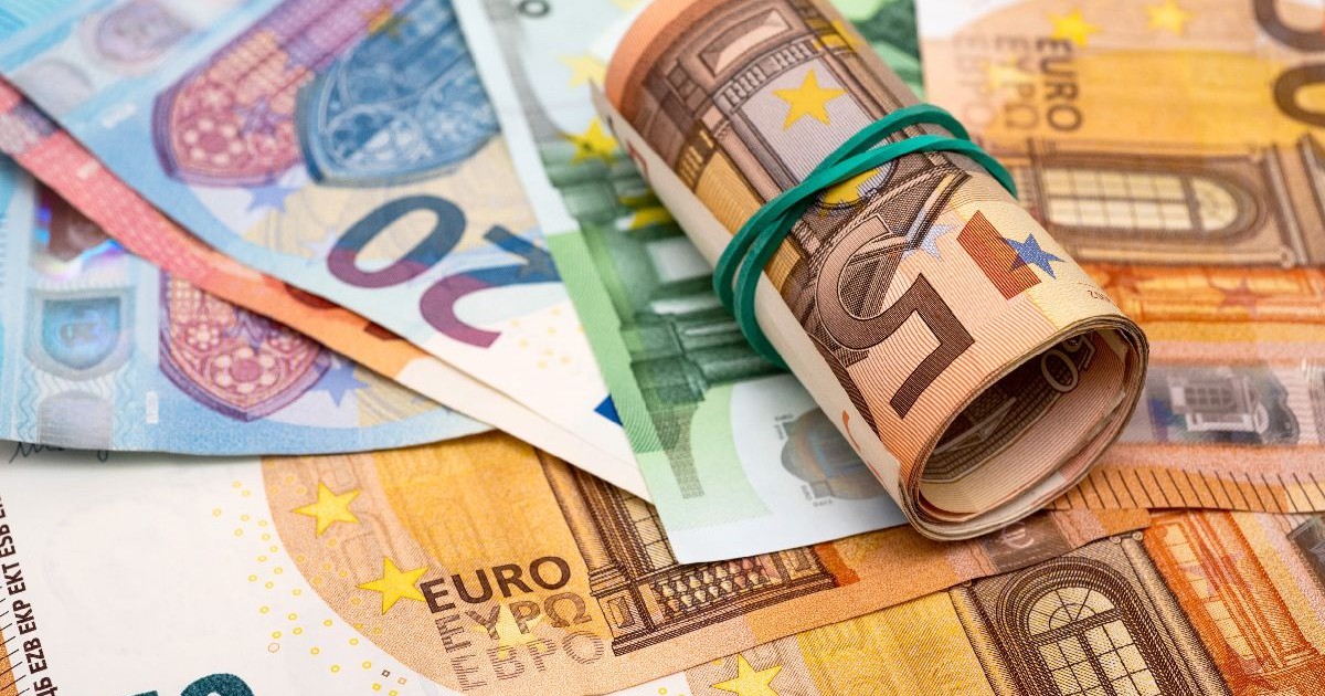 Imaginea prezintă bancnote euro cu valori diferite împrăștiate pe o masă, peste care este așezat un pachet de bancnote rulate și prinse cu un elastic de cauciuc verde.