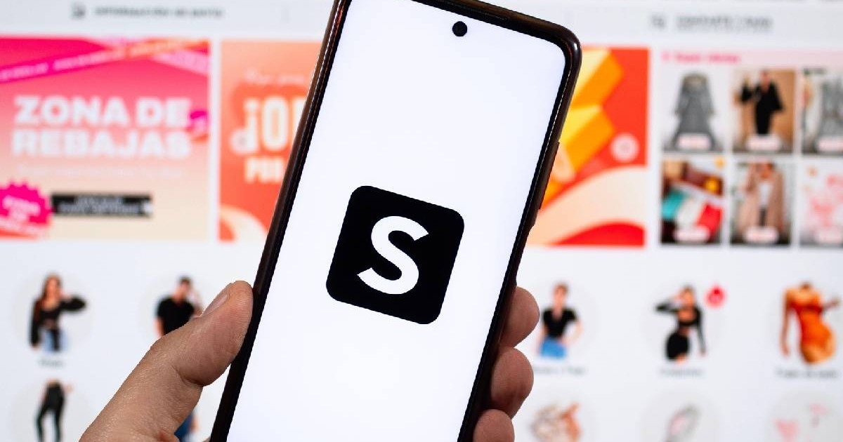 O mână masculină ținând un smartphone cu mâna stângă și simbolul Shein și o interfață Shein neclară