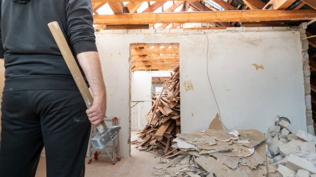 Un bărbat fotografiat cu spatele, îmbrăcat în negru, și cu un ciocan în mână este într-o casă în care e demolată o parte din ea