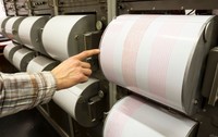 O mână care arată spre tamburul unui seismograf analogic pe care sunt înregistrate undele seismice