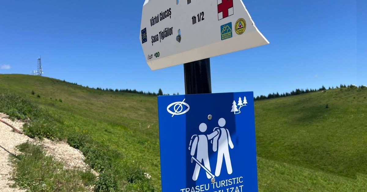 panou pătrat cu textul „TRASEU TURISTIC ACCESIBILIZAT” și pictogramele aferente, montat pe un stâlp metalic, chiar sub un indicator rutier clasic în formă de săgeată pe care scrie „Vârful Ciucaș” și „Șaua Țigăilor” alături de timpi estimați, panoul având ca fundal o culme montană înverzită pe care este amplasat un turn de telecomunicații.