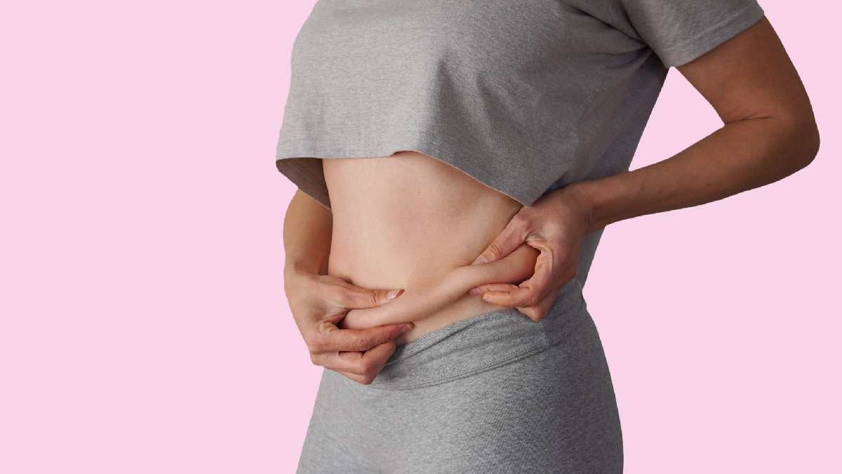 O femeie îmbrăcată în gri care strânge cu mâinile grăsimea abdominală.