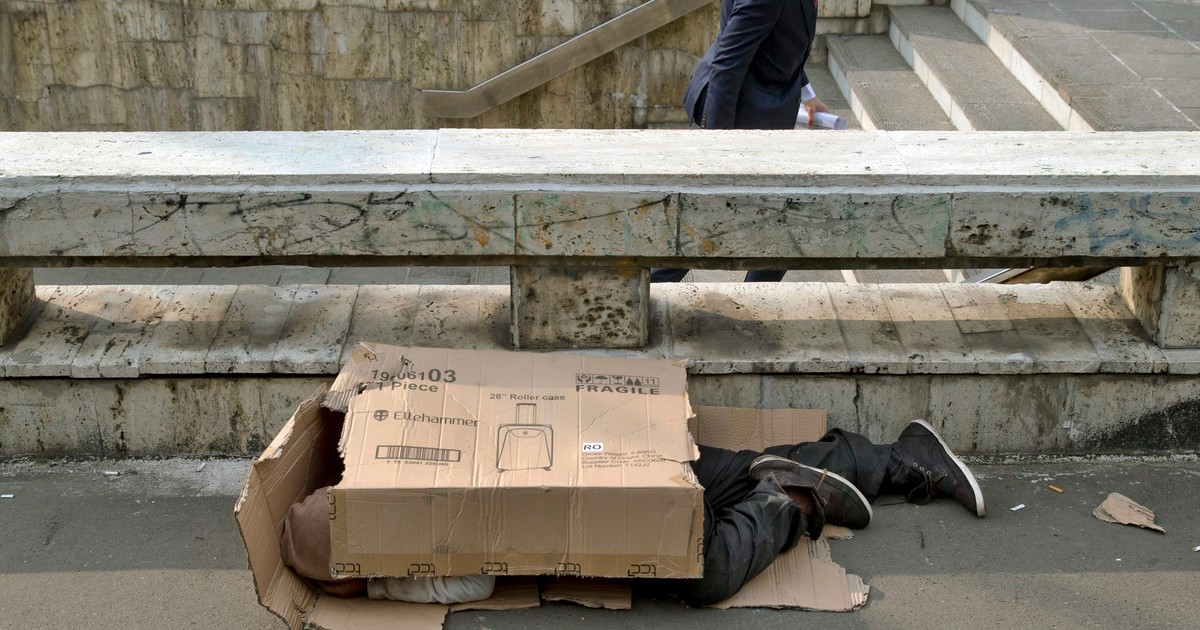 Un pieton iese dintr-o stație de metrou în timp ce un om fără adăpost doarme acoperit în ambalaje de carton, în fața gării principale din București, România