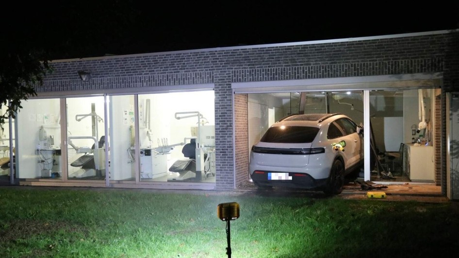 Un SUV Porsche alb care a intrat prin peretele de sticlă al unei clădiri moderne în care funcționează un cabinet stomatologic, pe timp de noapte.