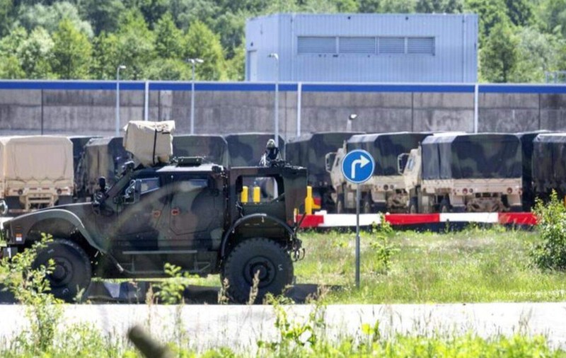 Un vehicul blindat de teren în prim-plan și o serie de camioane militare de transport aliniate în fața unui zid de securitate al unei baze militare.