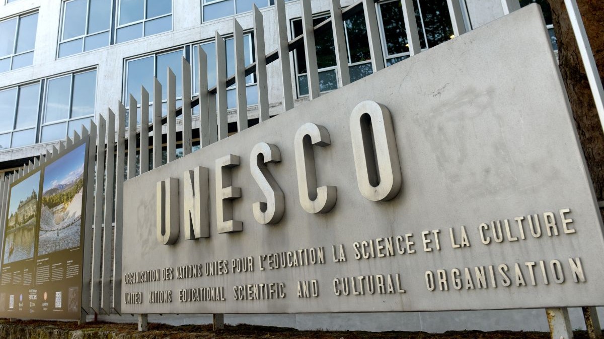 semn mare din piatră sau beton, pe care este scris cu litere mari, volumetrice, cuvântul „UNESCO”. Sub acest titlu apar texte gravate în franceză și engleză, descriind numele complet al organizației. Semnul este amplasat lângă un gard metalic, format din bare verticale lungi, iar în fundal se vede o clădire modernă cu multe ferestre mari, dispuse pe mai multe nivele.