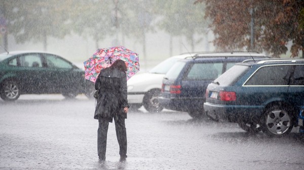 Imaginea prezintă o persoană îmbrăcată în haine închise la culoare care merge sub o umbrelă colorată în timpul unei ploi torențiale, într-o zonă de parcare cu autoturisme.   