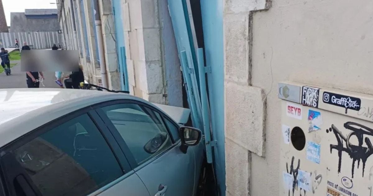 un autoturism Renault Clio de culoare gri argintiu, a cărui parte frontală este poziționată într-o deschizătură creată printr-o ușă metalică mare, de culoare bleu, care este înclinată și parțial desprinsă din structura unei clădiri cu pereți din piatră deschisă la culoare. În partea dreaptă a cadrului, pe zidul clădirii, se află un dulap metalic alb pentru utilități, acoperit de numeroase stickere și marcaje tip graffiti cu negru, în limba franceză, Châtellerault, Franța, 1 martie 2026
