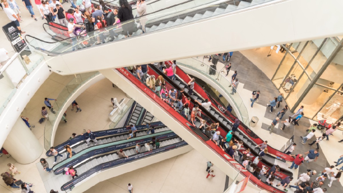 Vedere aeriană asupra interiorului aglomerat al unui mall, unde numeroase persoane utilizează scările rulante ce se intersectează la etaje diferite.