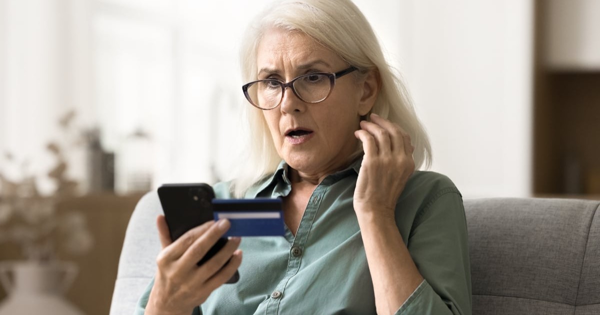 O femeie blondă cu ochelari care stă pe o canapea. Aceasta ține un telefon în mână, un card și este surprinsă.