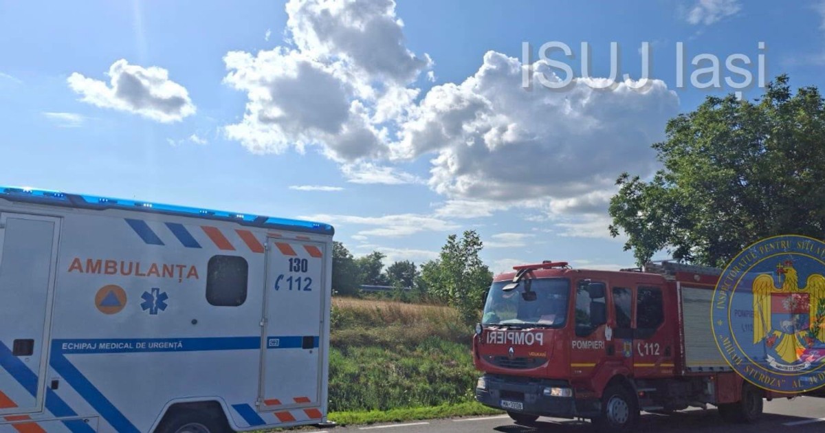 În imagine se observă o ambulanță și o autospecială de pompieri mobilizate la locul intervenției, pe marginea unui drum din județul Iași. Vehiculele de intervenție sunt staționate în apropierea zonei unde s-a produs accidentul feroviar, iar cerul senin contrastează cu gravitatea evenimentului.
