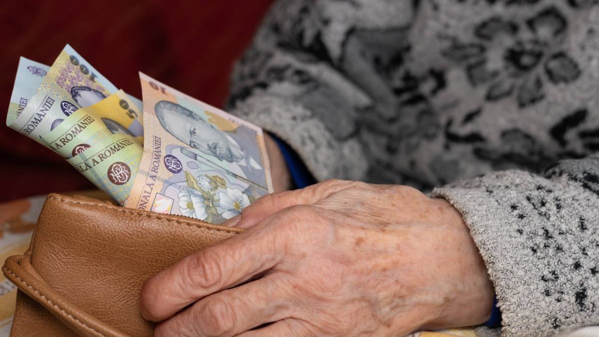 O femeie în vârstă care stă jos și își numără banii din portofel. Femeia poartă un pulover gri cu un model, portofelul pe care îl ține în mână este din piele maro, iar din portofel ies bancnote de 100, 50 și 10 lei.