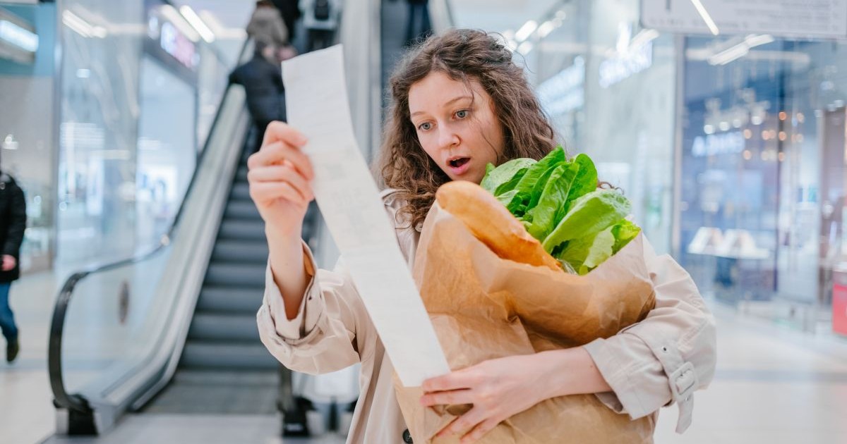 O femeie se uită foarte mirată la un bon de cumpărături într-un supermarket dintr-un mall și are în brațe un pachet cu pâine și salată, iar în fundal se văd scări rulante