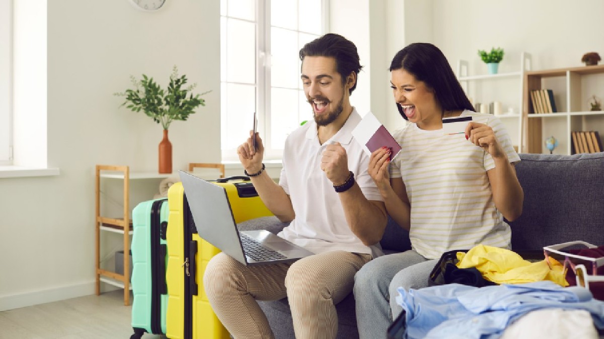 Cuplu fericit care se pregătește pentru o vacanță și plătește online . Un bărbat și o femeie, entuziasmați, stau pe canapea cu laptopul, cardul de credit, pașapoartele, biletele de avion și valiza de călătorie.