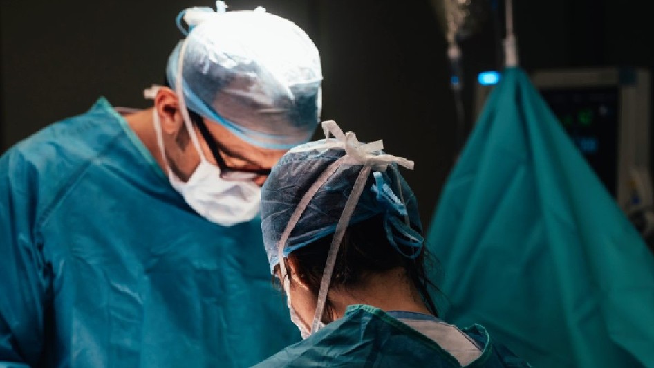 Doi medici, costumati in halate cu bonete si masti, care opereaza in sala de operatie