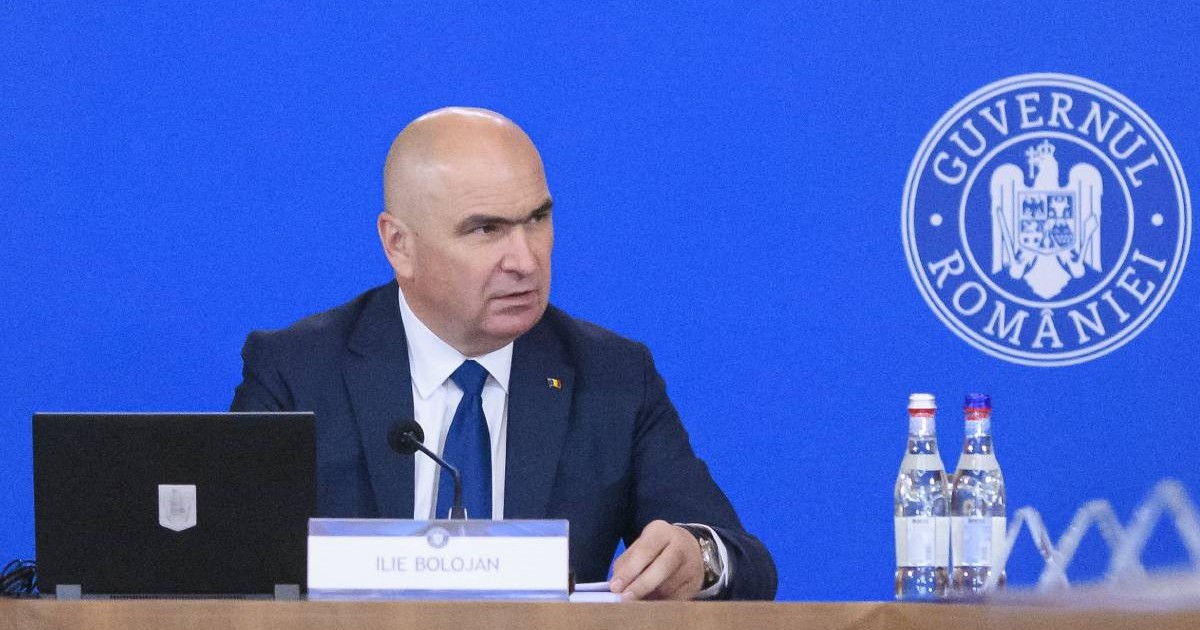 Ilie Bolojan la începutul ședinței de guvern, este îmbrăcat în costum și se uită în partea stângă.