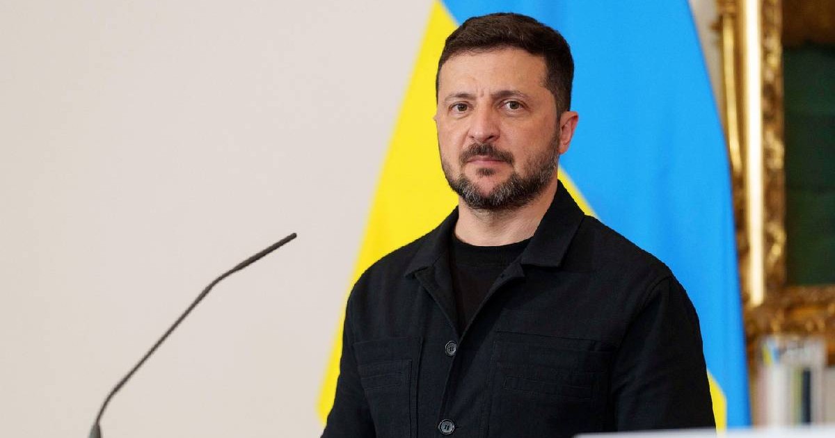 Volodimir Zelenski în timpul unei conferințe de presă, îmbărcat în negru, pe fundal se vede steagul Ucrainei