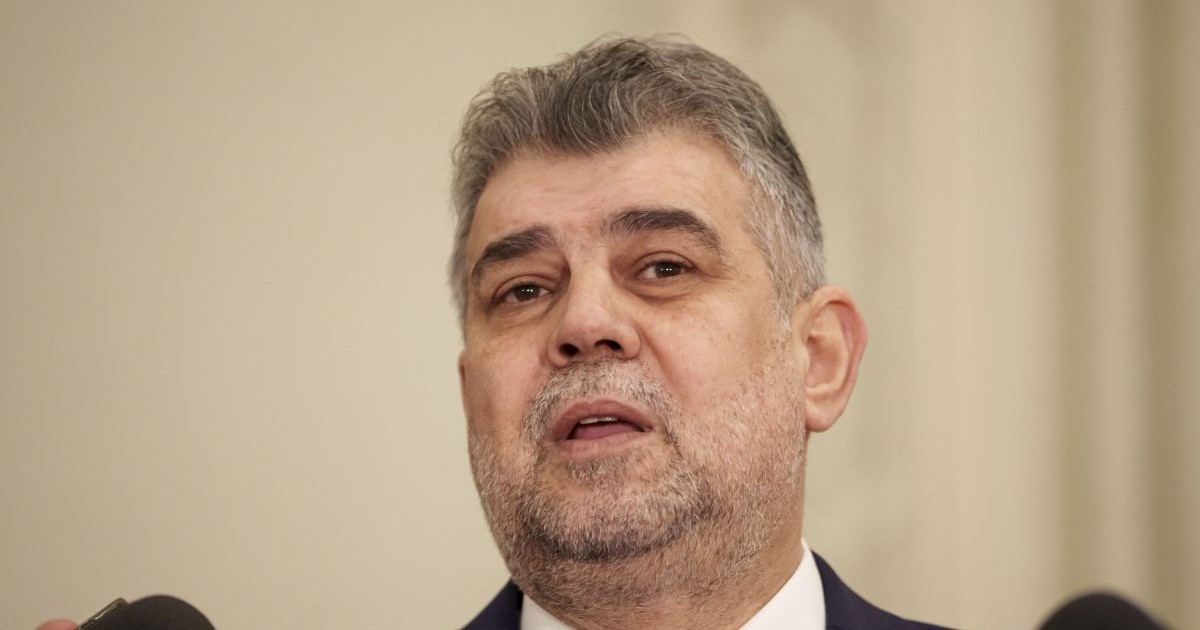 Marcel Ciolacu, în prim plan, îmbrăcat în costum cu cămașă albă, iar pe fundal un perete de culoare crem