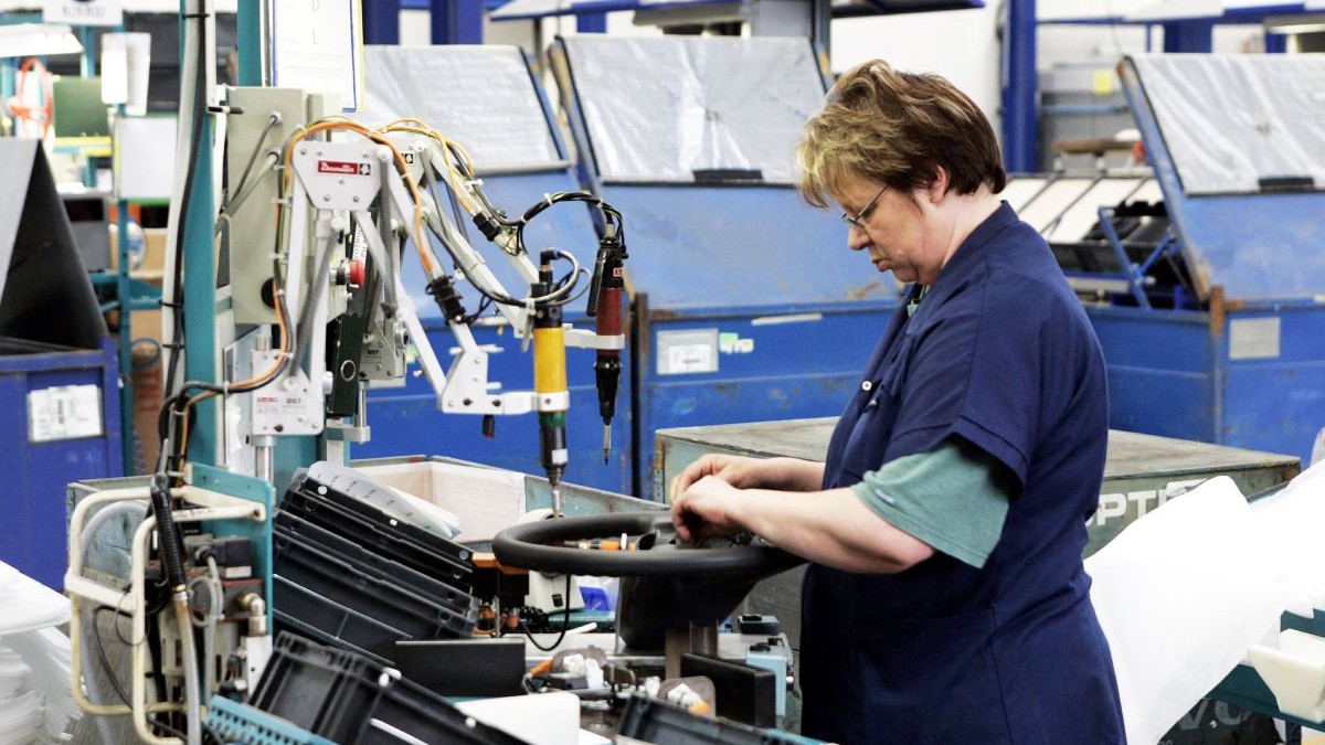 Imaginea prezintă o angajată care lucrează la asamblarea unui volan auto într-o fabrică industrială dotată cu echipamente tehnice specializate.