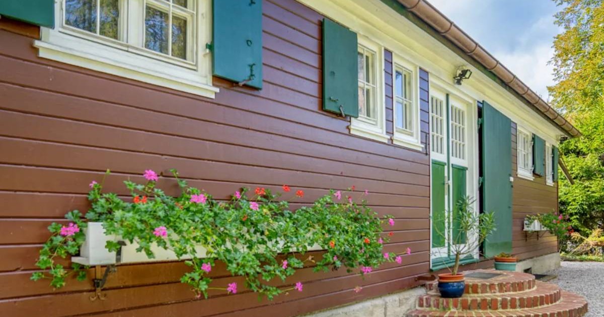 Casă din lemn, vopsită maro, cu obloane verzi și ferestre albe, decorată cu flori colorate la exterior și o mică scară de cărămidă la intrare.