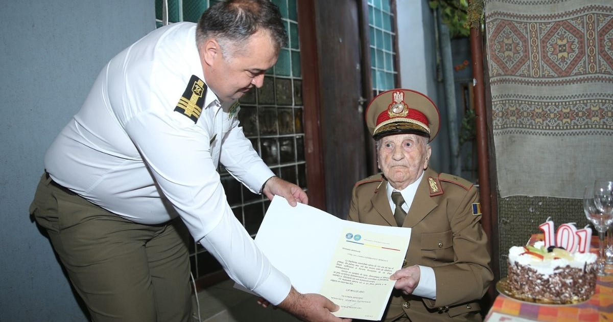 Veteranul de război Andrei Georgescu-Ionescu, la petrecerea de aniversare a vârstei de 101 ani, stând pe scaun îmbrăcat într-o uniformă militară lângă un tort cu candele cu cifra 101