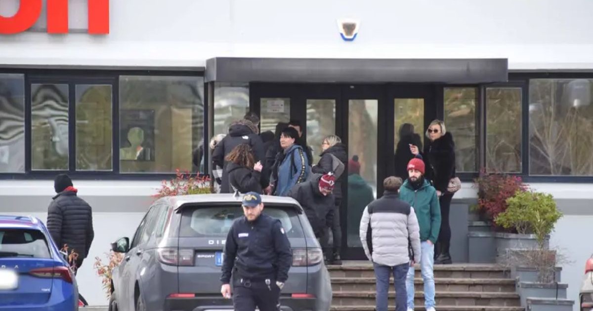 Un grup de persoane, printre care se află și un polițist, stau în fața unei clădiri, cu ferestre mari, așteptând la intrare.