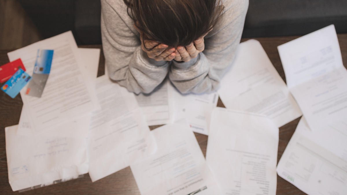 O persoană care își ține capul în mâini deasupra unei mese pline de facturi, documente împrăștiate și carduri bancare, sugerând o stare de stres sau epuizare financiară