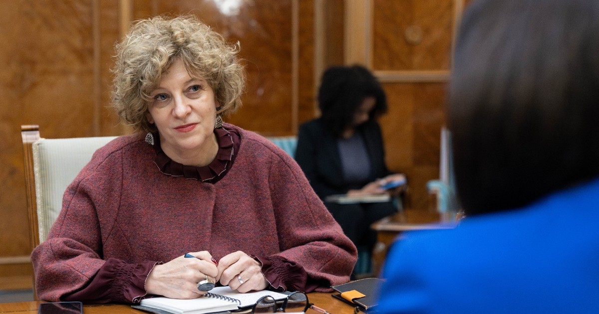 Vicepremierul Oana Gheorghiu, în timpul unei ședințe la Palatul Victoria, poartă un palton vișiniu și stă așezată pe scaun, la masă