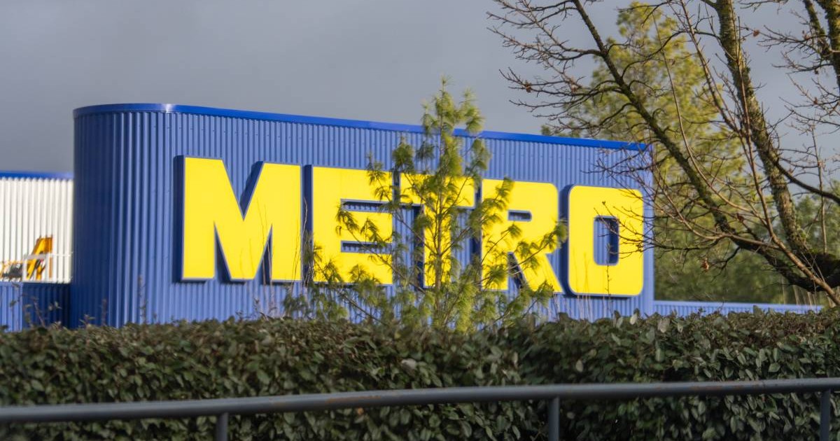 Sigla METRO, cu litere galbene strălucitoare pe clădirea albastră în spatele verdeții luxuriante