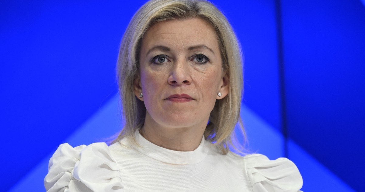Imaginea este un portret realizat pe un fundal albastru aprins, care o surprinde pe Maria Zaharova în prim-plan, având o expresie serioasă și privirea fixă. Ea poartă o bluză albă cu mâneci bufante și are părul blond, aranjat scurt.