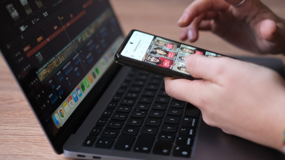 Mâinile unei persoane care utilizează un smartphone peste tastatura unui laptop, pe ecranul laptopului fiind deschis un program de editare video, iar pe ecranul telefonului o interfață cu conținut media.