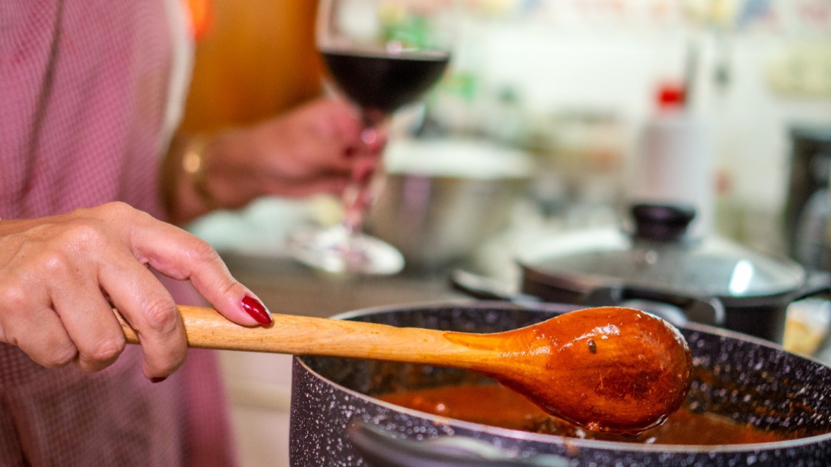 O femeie în bucătărie care gătește. În mâna stângă ține un pahar de vin