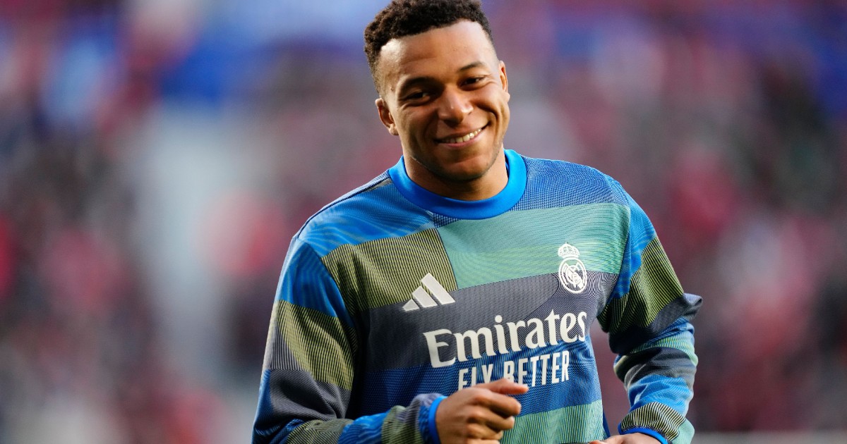 Mbappe zâmbește în timp ce face încălzirea pe teren, purtând un tricou de antrenament al clubului Real Madrid cu sponsorul „Emirates Fly Better”. În fundal se vede stadionul plin, iar atmosfera sugerează momentele dinaintea unui meci important.