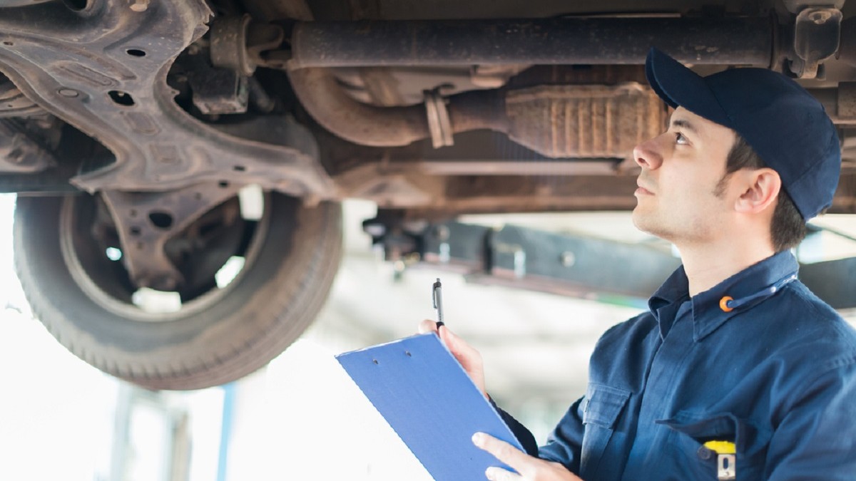 Ce este flitrul de particule al unei maşini şi cum se întreţine - Un mecanic auto verifică partea inferioară a unei masini, ridicată pe elevator, într-un service, ținând în mână un dosar clipboard cu documente pentru notițe