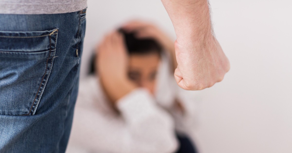 Imagine cu o femeie care stă pe podea și-și ține mâinile pe cap, iar în stânga se află un bărbat cu pumnul încleștat