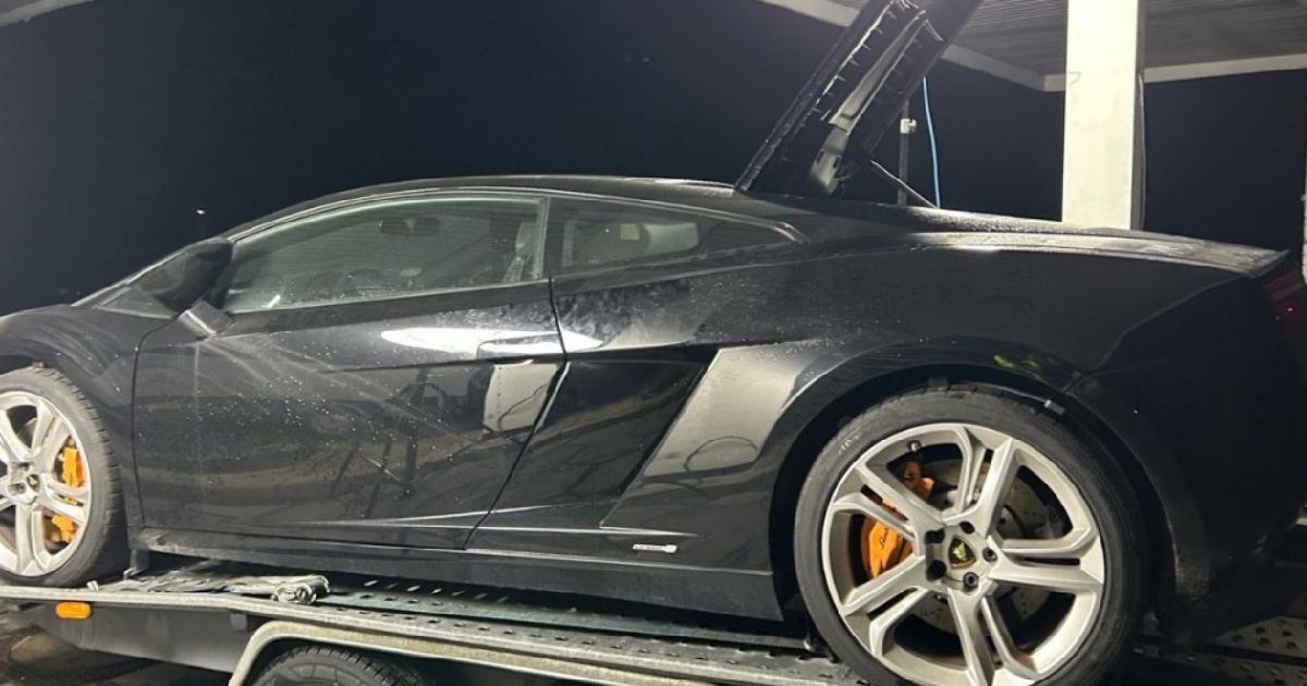 Imaginea prezintă un Lamborghini negru așezat pe o platformă. Mașina are jante mari din aliaj argintiu. Fundalul este întunecat.