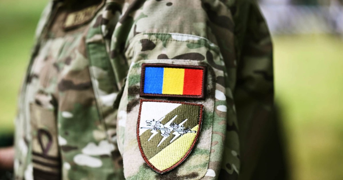 Imaginea prezintă un prim-plan al mânecii unei uniforme militare de camuflaj, pe care este cusut un ecuson cu drapelul României.