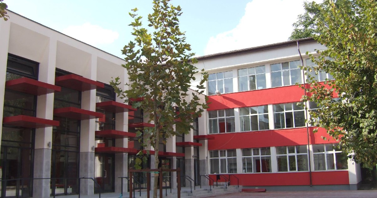 Imaginea prezintă exteriorul unei clădiri moderne, o școală, cu două corpuri distincte: unul de culoare albă, cu ferestre mari, și altul de un roșu intens. Curtea interioară este simplă, cu câțiva copaci tineri și o bancă, sugerând un spațiu curat și funcțional.