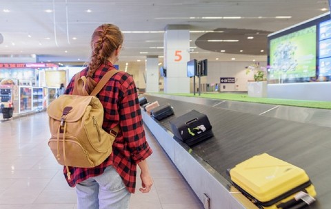 De ce să fotografiezi bagajul înainte de a pleca în vacanță - Imagine cu o femeie cu un rucsac în spate aşteptându-şi bagajul de cală la banda de bagaje dintr-un aeroport