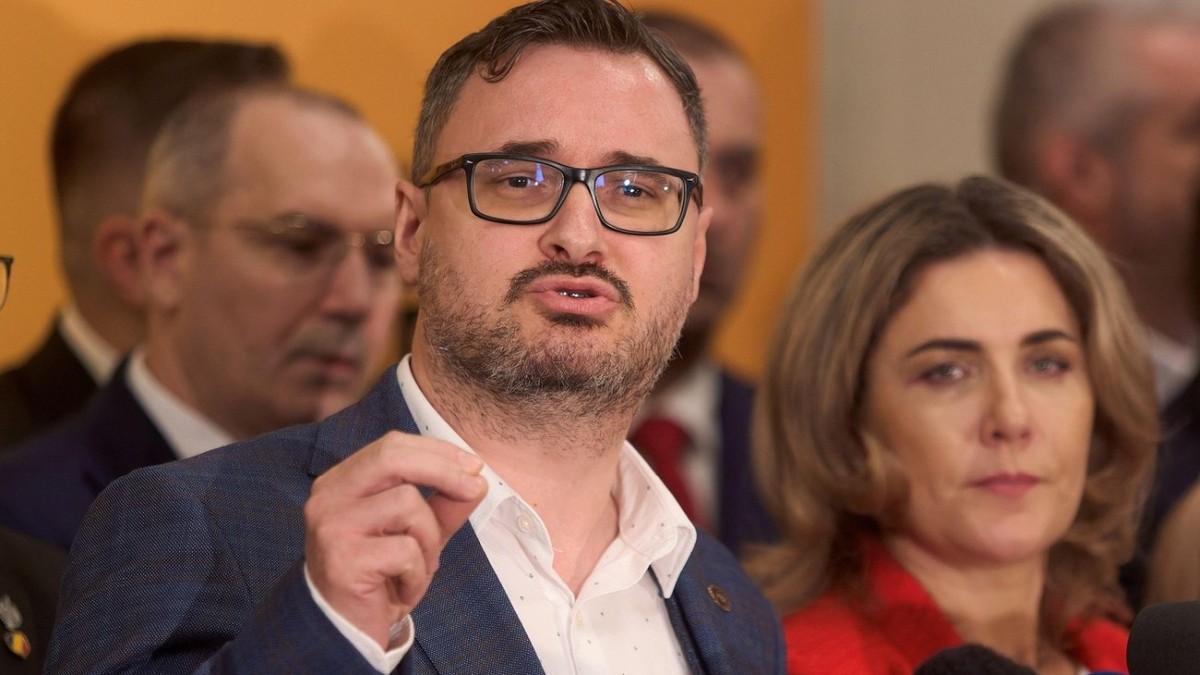 Dan Tanasă, intr-un sacou gri și o cămașă albă, vorbind și gesticulând, ținând o mână ridicată și atingându-și degetul arătător de cel mare
