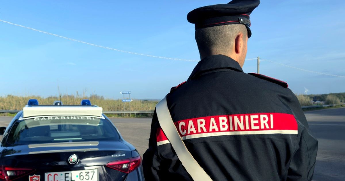 Fotografie a unui ofițer italian al Carabinieri în uniformă, cu spatele la cameră, stând lângă un sedan Alfa Romeo albastru parcat.