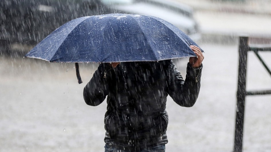 O persoană îmbrăcată într-o geacă neagră care se adăpostește sub o umbrelă albastră, ținând-o strâns cu ambele mâini în timpul unei ploi torențiale.