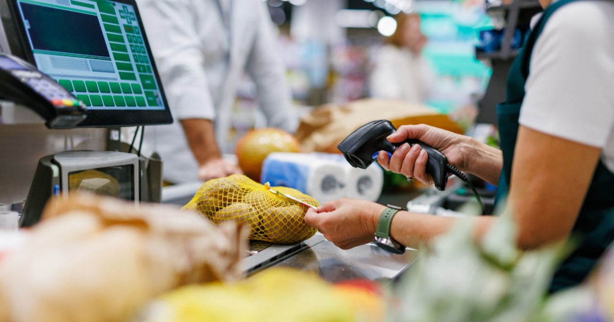 Imaginea prezintă în prim-plan mâinile unei casiere de supermarket în timp ce folosește un scanner portabil pentru a înregistra codul de bare al unei plase cu legume la casa de marcat.