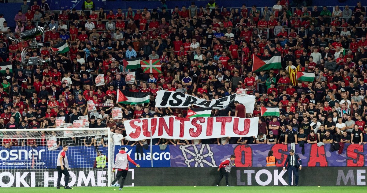 Imaginea surprinde un moment dintr-un meci de fotbal (posibil Osasuna-Getafe conform numelui fișierului), cu o tribună plină de suporteri îmbrăcați majoritar în roșu, care afișează diverse steaguri și bannere cu mesaje politice și locale. Printre steagurile palestiniene și basce, în centru se remarcă un banner mare cu textul "SUNTSITU" scris cu roșu pe fundal alb, sub un alt banner care pare să menționeze Israel.