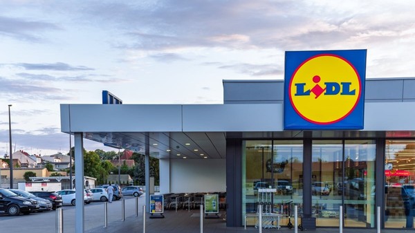 Un magazin Lidl modern este surprins din exterior, cu intrarea largă și logo-ul vizibil deasupra ușilor automate. În fața clădirii se află parcarea și zona pentru cărucioare, sugerând acces facil pentru clienți.