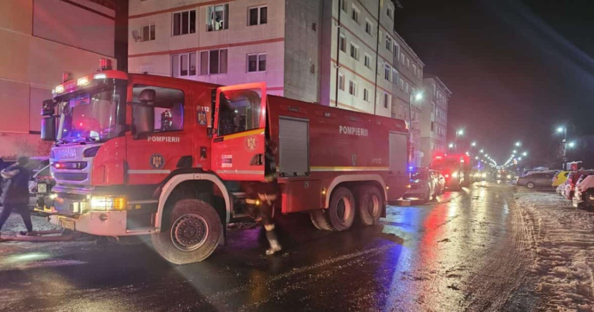 Imaginea surprinde o intervenție a pompierilor pe timpul nopții, cu autospeciale de stingere și lumini de avertizare aprinse, în fața unui bloc de locuințe. Carosabilul umed și prezența echipajelor de intervenție sugerează o situație de urgență recentă, cel mai probabil un incendiu.