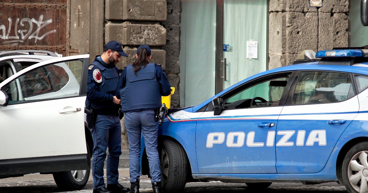 Imaginea surprinde doi ofițeri de poliție, un bărbat și o femeie, stând lângă o mașină de poliție albastră marcată vizibil cu cuvântul "POLIZIA". Aceștia par să fie angajați într-o discuție sau să inspecteze un incident , având o mașină civilă albă parcată lângă vehiculul lor de serviciu.