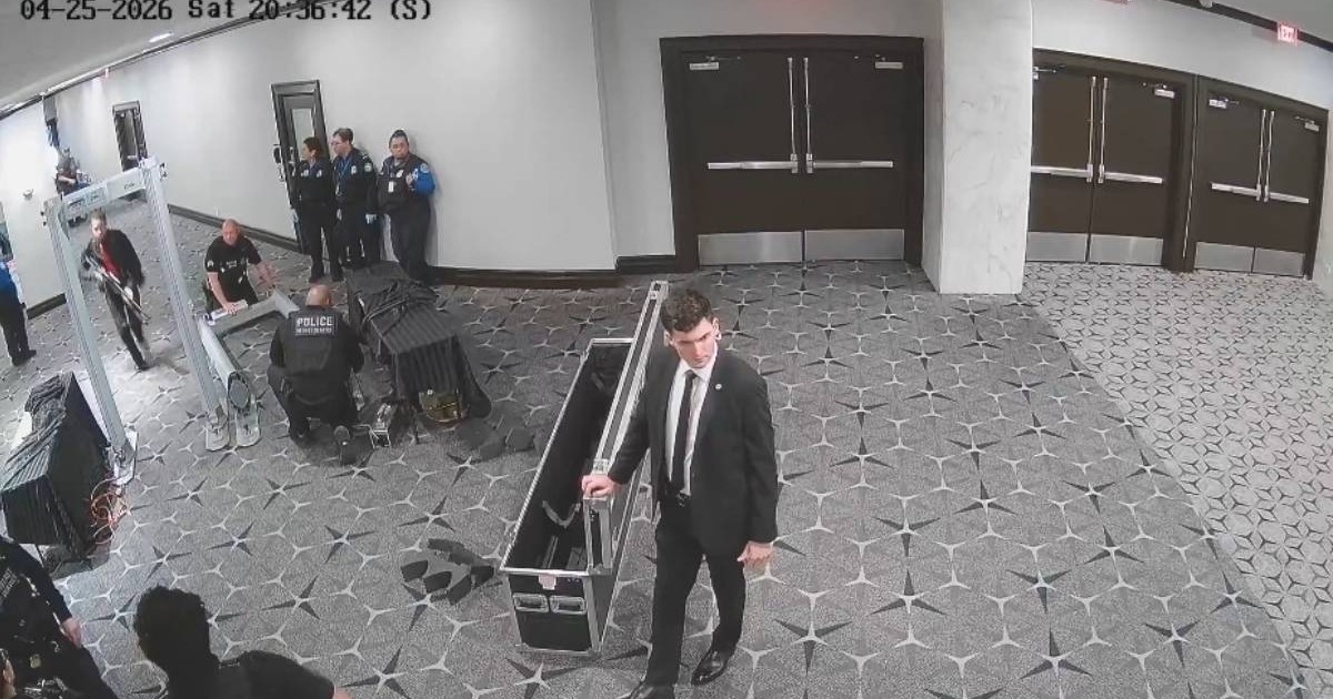 Imagine surprinsă de o cameră de supraveghere într-un hol al hotelului Washington Hilton, pe 25 aprilie 2026. În cadru se vede un bărbat îmbrăcat la costum, lângă o cutie mare de transport deschisă, în apropierea unui filtru de securitate. În stânga imaginii sunt mai mulți agenți de pază și polițiști, iar în fundal se văd uși mari închise și holul hotelului.