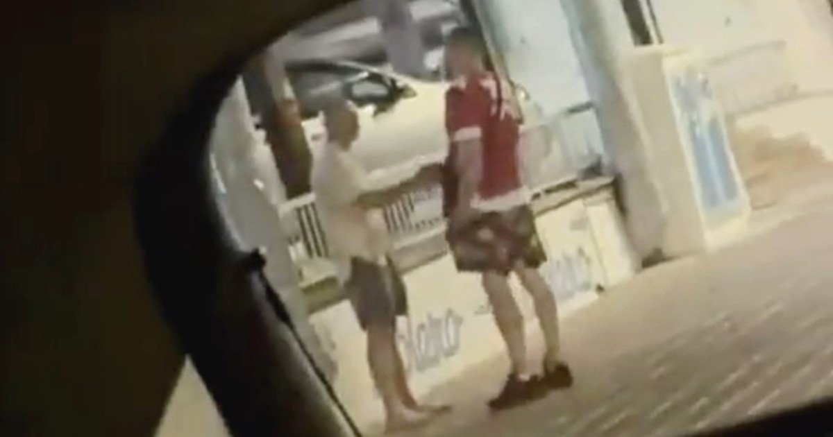 Imaginea prezintă doi bărbați care discută pe un trotuar pe timp de noapte, surprinși prin geamul sau oglinda unui vehicul.