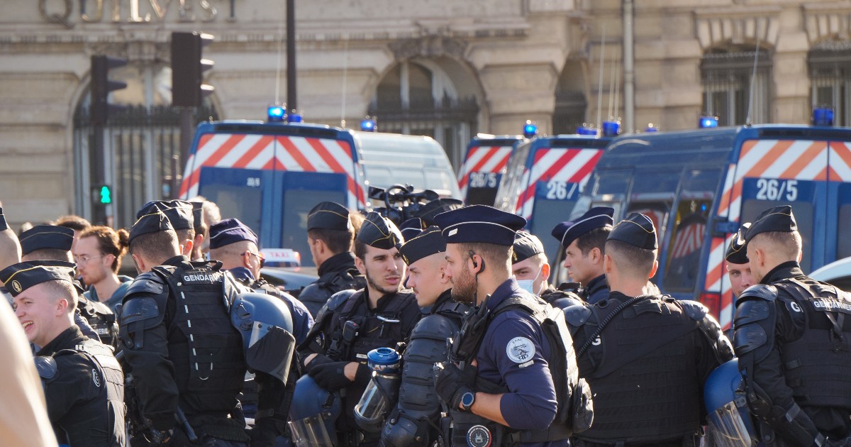 Imaginea arată un grup de ofițeri de poliție și jandarmi îmbrăcați în uniforme de culoare închisă, cu veste de protecție și căști. Unii dintre ei poartă berete negre, iar unii par să aibă părul tuns scurt. O parte din ofițeri stau de vorbă între ei. În fundal se pot vedea mai multe dube de poliție albastre, cu dungi roșii și albe, sugerând că se află în timpul unei misiuni sau că patrulează.